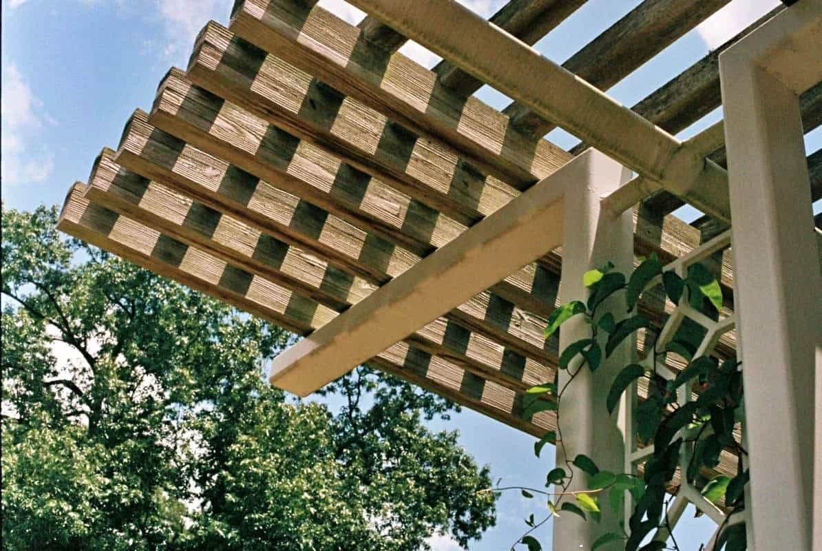 Dřevěná pergola s mřížkovou střechou pod modrou oblohou.