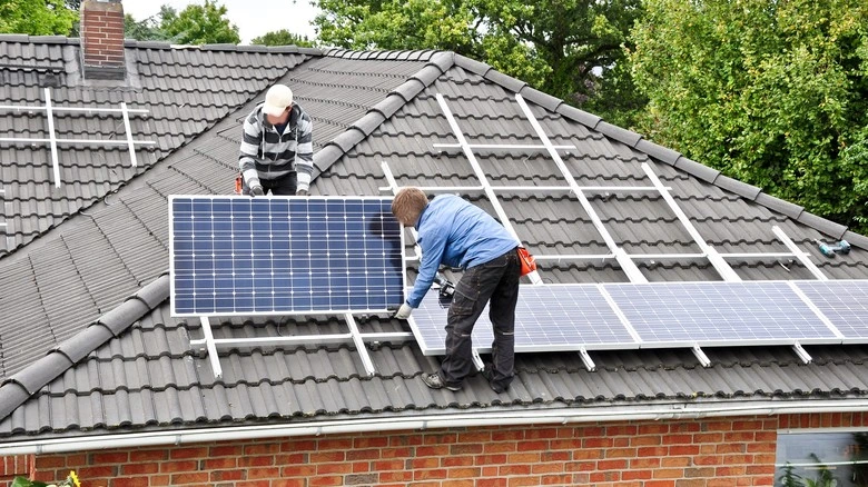 Dva dělníci montují fotovoltaické panely na střechu domu.