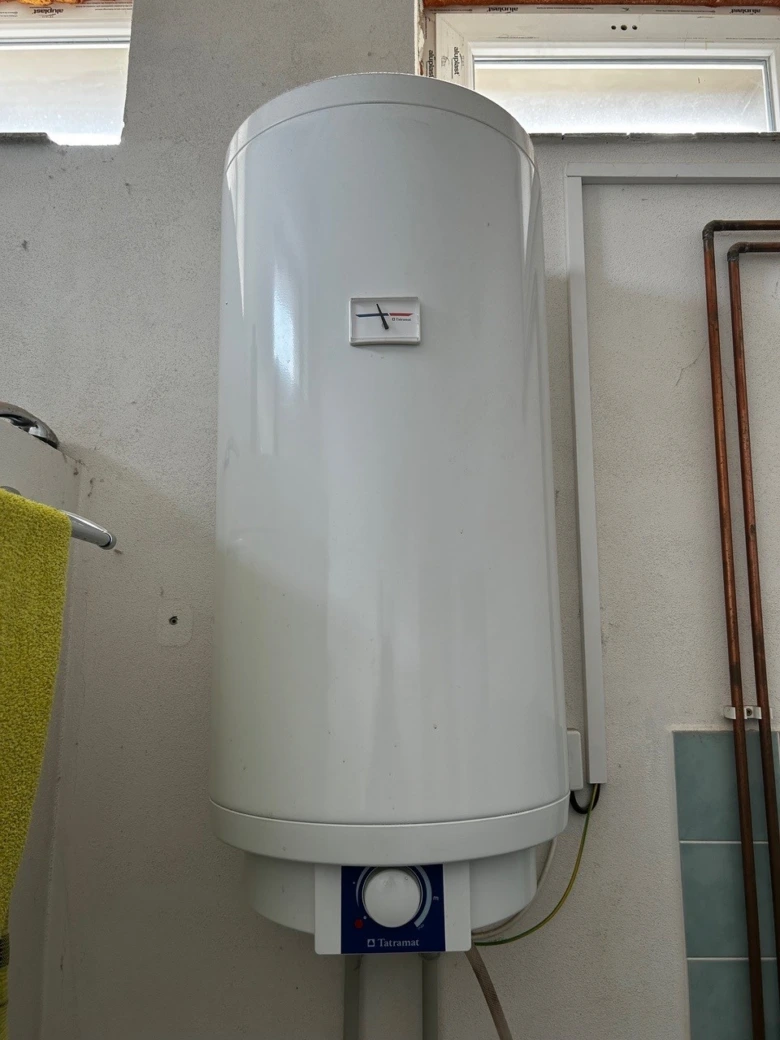 Bílý elektrický bojler na zdi s viditelnými potrubím.