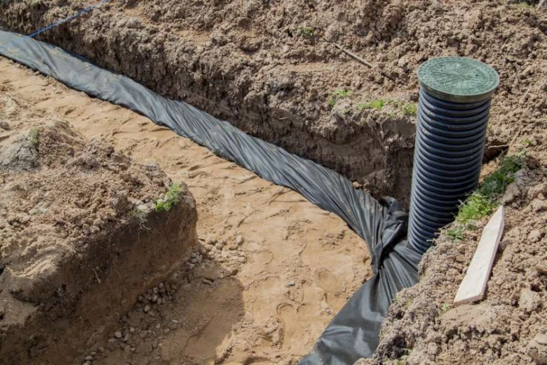 Instalace černé drenážní trubky v příkopu s geotexilem.