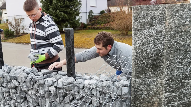Muži plní drátěný koš kameny pro stavbu gabiónové zdi.