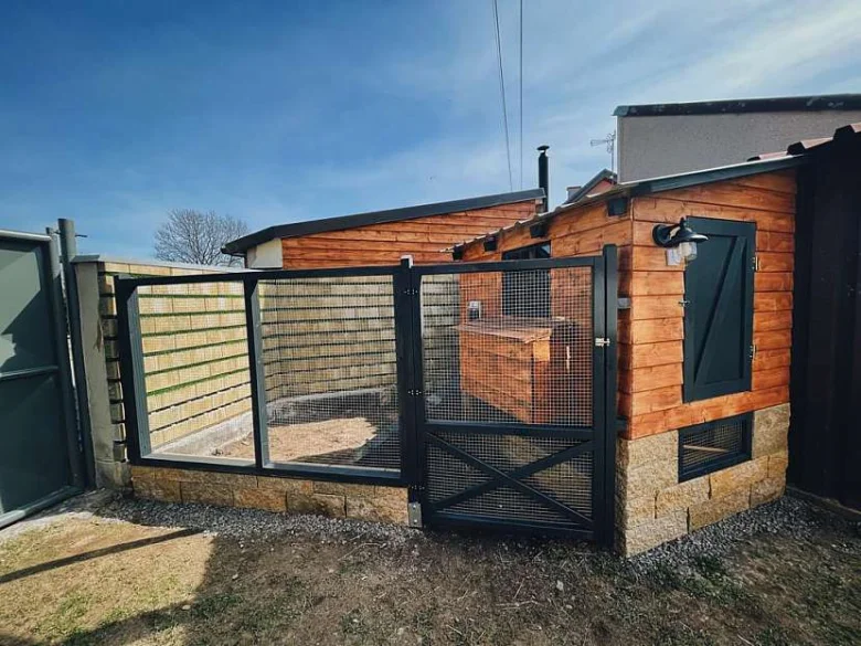 Hotový dřevěný kurník s kamenným základem a kovovým plotem.