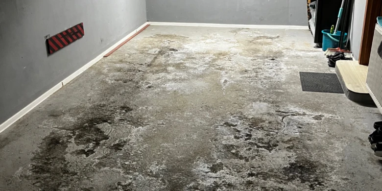 Poškozená betonová podlaha v garáži s plísní a skvrnami.