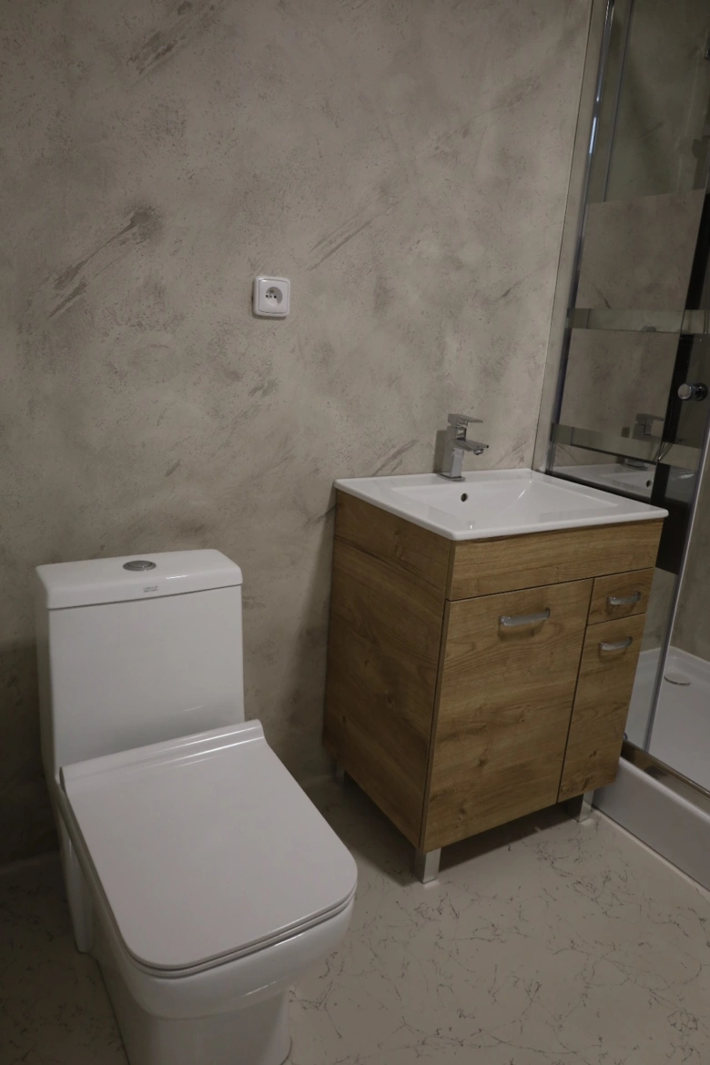 Koupelna s betonovou stěrkou na zdi vedle WC a umyvadla.