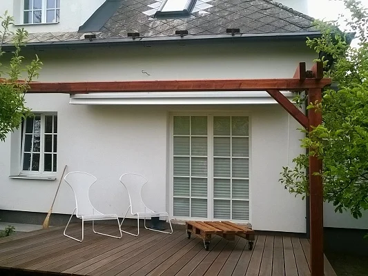 Dřevěná podlaha na terase pod pergolou u bílého domu.