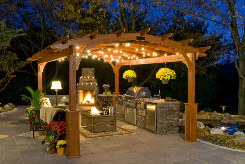 Osvětlená dřevěná pergola s krbem a grilovnou na zahradní terase večer.