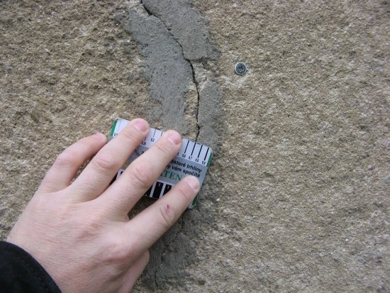 Ruka měří šířku trhliny v betonu při kontrole statiky domu.