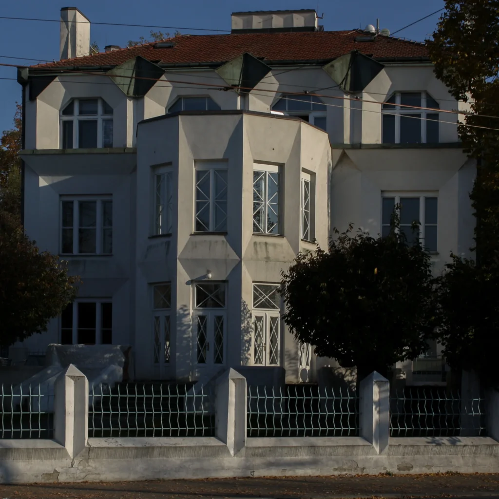Bílá kubistická vila s geometrickými okny za plotem.