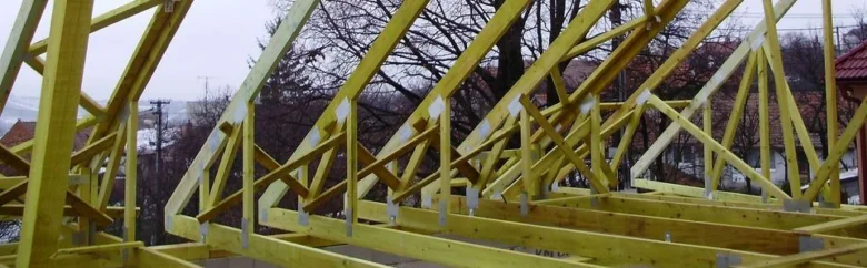 Žluté dřevěné vazníky tvořící střešní konstrukci na stavbě.