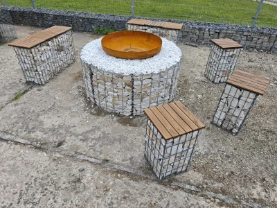 Kruhový ohniště z gabiónů s sedacími lavičkami na štěrkové ploše.