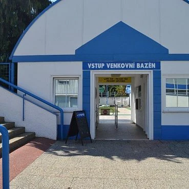 Budova vstupu venkovního bazénu s modrým štítem a schody vedoucí dovnitř.