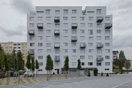 Moderní bílá panelová budova s balkóny před řadou mladých stromů.