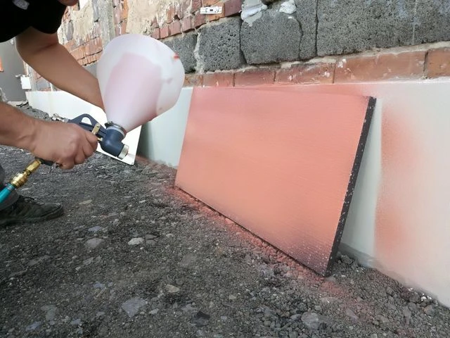 Pracovník aplikuje růžovou fasádní omítku na zdivo pomocí spreje.