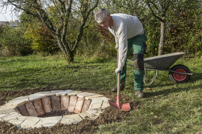Muž staví ohniště z cihel na zahradě lopatou.