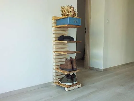 Dřevěný botník s obuví, krabicí a dekorací na bílé stěně.