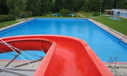 Červená vodní skluzavka do bazénu v koupališti s okolním trávníkem.